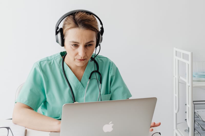 Healthcare professional in scrubs listening intently through headphones at a laptop, working through the 90 second preparation window in OET Listening Part C.