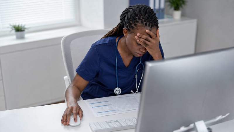 A healthcare professional at a clinic desk, head in hand, in front of a practice test on screen: the outdated mock trap, scoring well at home and failing the real exam.