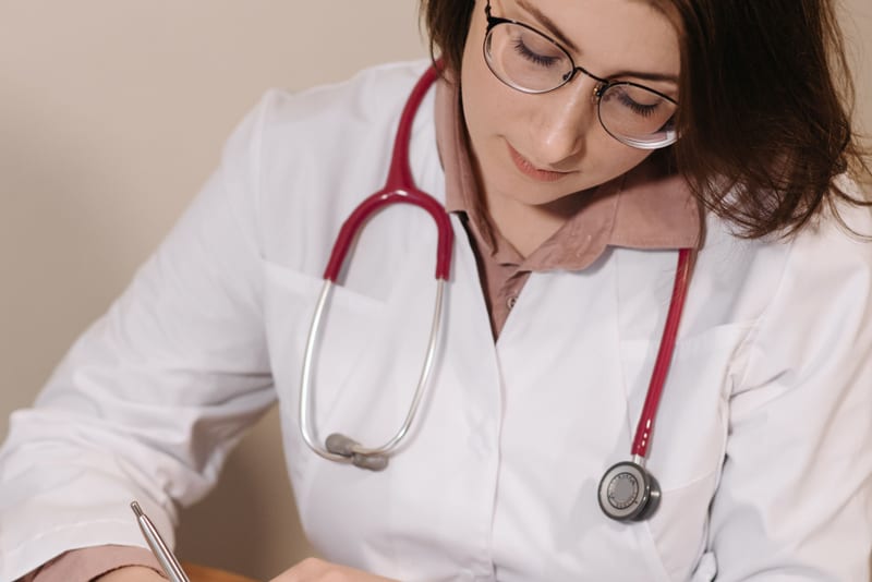 Doctor in a white coat writing a referral letter by hand, building the story before the letter goes on the page.